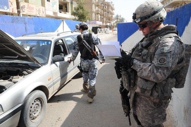 BAGHDAD -  A National Policeman searches a car at a checkpoint while Staff Sgt. Joshua Bell of Battery A, 1st Battalion, 319th Airborne Field Artillery Regiment,  takes notes on his clipboard during an inspection of Iraqi Security Forces...