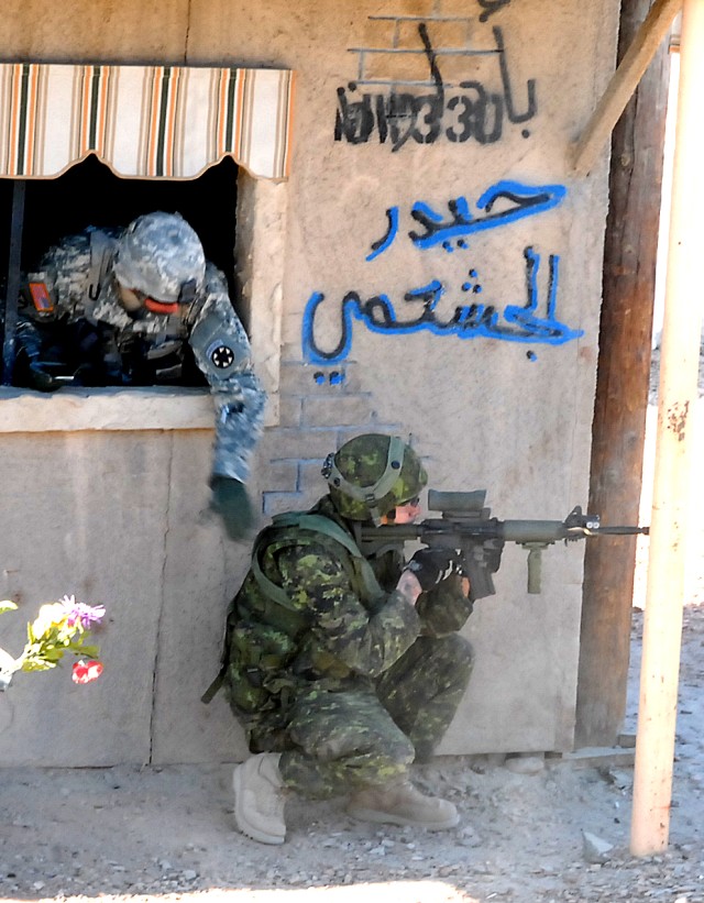 A Canadian Soldier positions himself at the corner of a building in the village of Medina Wasl at the National Training Center at Fort Irwin, Calif., as an American observer-controller with Operations Group taps his shoulder during a training exercis...
