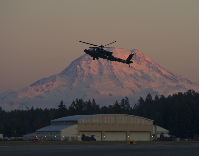 An AH-64E Apache flies over a hangar with snow-covered mountains in the background. The 1-229th Attack Reconnaissance Battalion at JBLM became the first unit equipped with the E model Apache in May 2013.