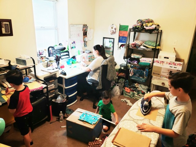 Courtney Bell works at her sewing machine as her children form an assembly line to feed her the pieces she needs to make masks. Antonio, 9, (right) irons mask fabric squares for Gabriel, 5, (left) who takes them and traces the mask outline on the...