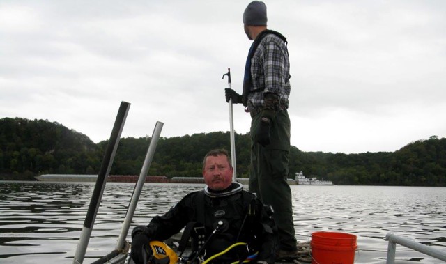 A standby diver and tender wait on board a dive vessel with barge traffic in the background.  Divers use dive helmets and surface supplied air during this operation.