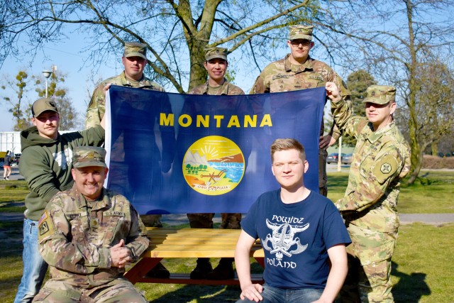 Clockwise from left, seated: Maj. Colin Curry, Spc. Dylan Roselles, Maj. Brian Casey, 1st Sgt. Alicia Roethler, Sgt. Colton Smith, Sgt. 1st Class John Proulx, and Pfc. Koleton Grimes, all Soldiers from the 652nd Regional Support Group out of Helena, Montana, pose with the Montana state flag April 6, 2020 on “406” day in Powidz, Poland. The Montana Soldiers with the 652nd RSG are wrapping up a nearly year-long deployment to Poland by quarantining at North Fort Hood, Texas. (U.S. Army Reserve photo by Maj. Olha Vandergriff, 652nd Regional Support Group)