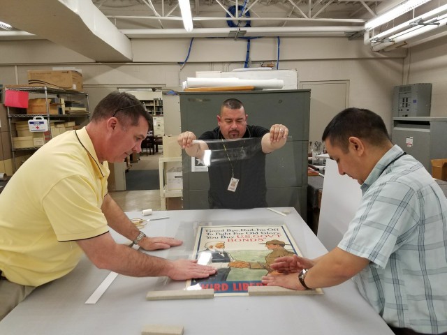 Museum Specialist Chris Goodrow, Exhibit Specialist Lupe Perez, and Archivist Carlos Alvarado work on the finishing touches for featured posters on display in the WWI Gallery Exhibit at the U.S. Army Medical Department Museum. Photo courtesy of...