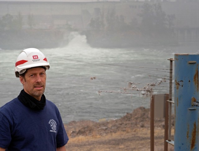 Jim Day, fisheries biological technician inspects avian wires at The Dalles Lock and Dam-seen in the background. The wires help deter birds from preying on juvenile salmon as they migrate downstream through the sluiceway.