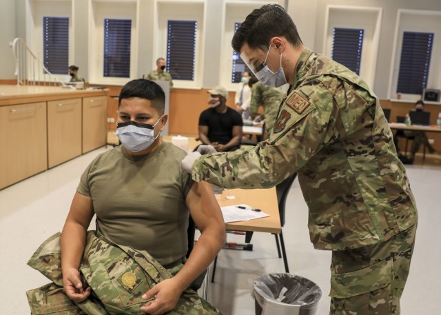 U.S. Army 1st Lt. Fabian Zuniga (left), a staff nurse at Landstuhl Regional Medical Center, receives the first inoculation of the Moderna COVID-19 vaccine administered by U.S. Air Force Airman 1st Class Mike Leontyuk (right), a medical technician...