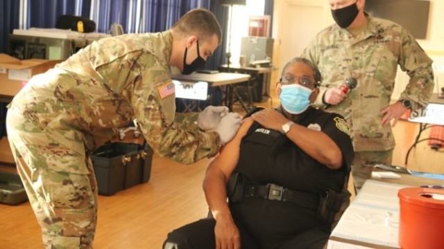 Security guard Joseph Butler III, with the Directorate of Operations, receives the first COVID-19 vaccination on post from Sgt. Michael Corral, with the U.S. Army Medical Research Institute of Chemical Defense, while APG Garrison Commander Col. Timothy Druell looks on during a Facebook broadcast at the APG North (Aberdeen) recreation center Jan. 7. 