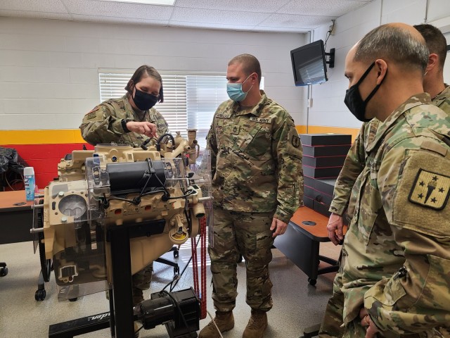 Sgt. 1st Class Ricardo Arbelaez (center) demonstrates the engine training device used during basic and advanced level Wheeled Vehicle Mechanic (91B10/30) courses to Command Sgt. Maj. Petra M. Casarez (left) and Chief Warrant Officer 5 Danny K. Taylor (right).