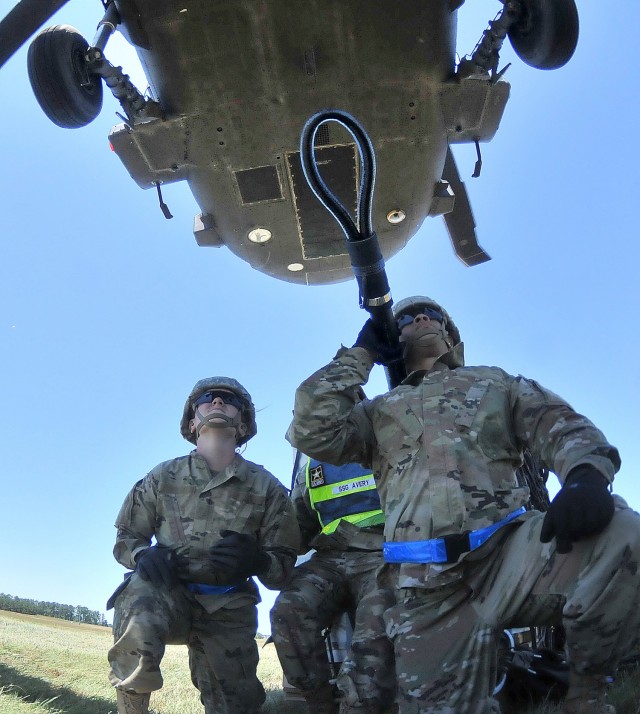 A Black Hawk helicopter slowly hovers downward toward students and instructor,  facilitating the task of using the reach pendant to connect cargo to the aircraft for transport during sling load training June 17 at McLaney Drop Zone. The  training event was part of the 89B Ammunition Specialist Course taught at the Ordnance School.