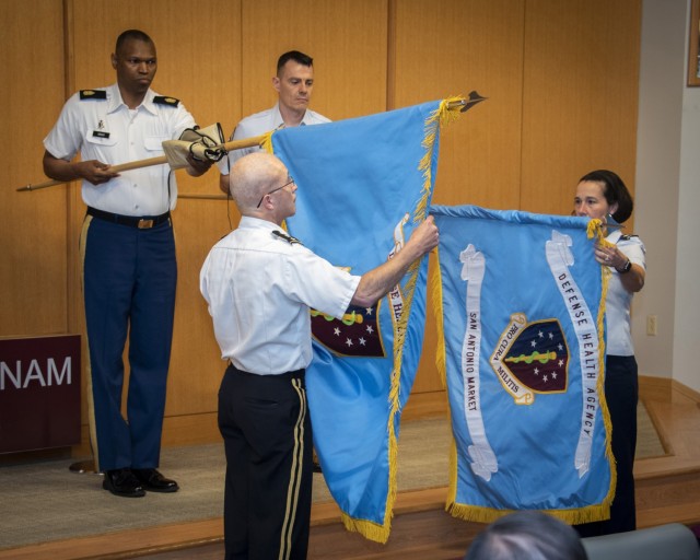 Army Lt. Gen. Ronald J. Place, director of the Defense Health Agency, unfurls the DHA flag as Air Force Brig. Gen. Jeannine M. Ryder, 59th Medical Wing commander and San Antonio Market director, unveils the new San Antonio Market flag during the market establishment ceremony at Brooke Army Medical Center, Fort Sam Houston, Texas, July 16, 2021. Army Command Sgt. Maj. Michael L. Gragg, Defense Health Agency senior enlisted leader, and Air Force Chief Master Sgt. Marc Schoellkopf assist in unveiling the flags. (U.S. Army photo by Jason W. Edwards)