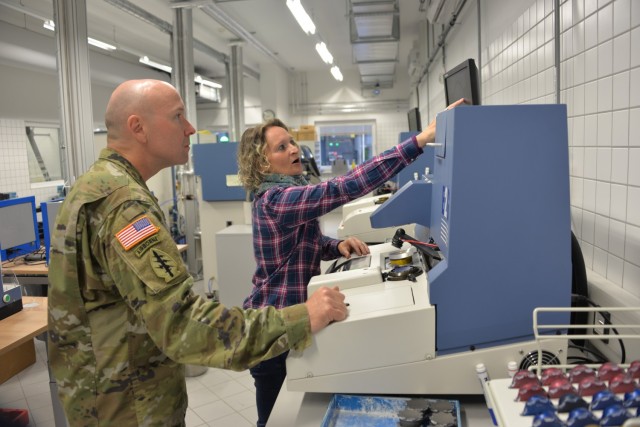 Yvonne Gortner, right, prescription eyeglass maker supervisor for USAMMCE works with Col. Shane Roach, USAMMC-E commander, to grind a new pair of custom eyeglasses. The digitally controlled equipment grinds a lens from a blank lens to create custom lenses for non-typical prescriptions.