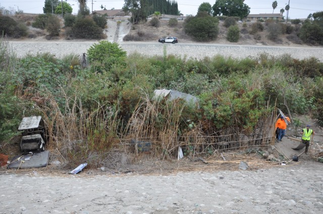 Contracted workers begin hauling debris up the steep levee bank as a Los Angeles County Sheriff patrol maintains safety. Note the use of Arundo donax, a pernicious invasive grass resembling bamboo that can grow a foot or more per day, for fencing....