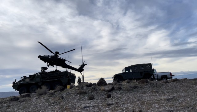 An AH-64 Apache flies over a Stryker assigned to 2nd Stryker Combat Team, 7th Infantry Division and a Toyota Mega Cruiser assigned to the Japanese Ground Self-Defense Force during training exercise Rising Thunder on December 6, 2021 at Yakima Training Center, Wa.