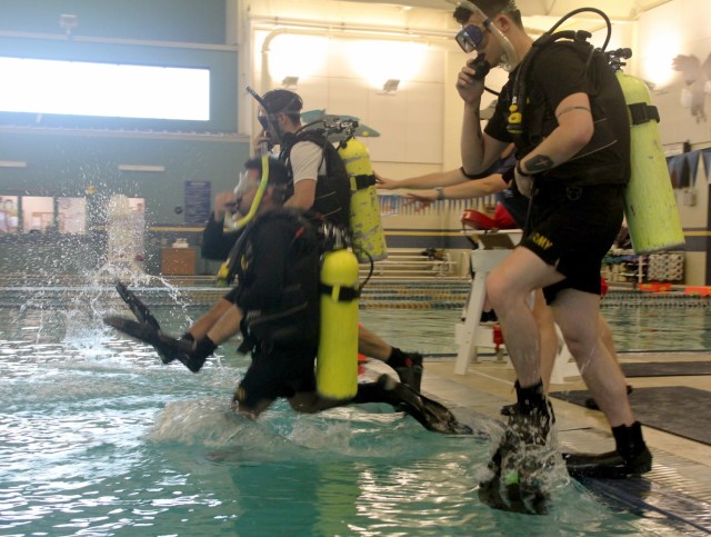 A group of students, some wearing dive equipment for the first time, jump into the pool at Naval Station Everett, Washington, during the Joint Hyperbaric Medical Officer and Technician Course, April 26, 2022.