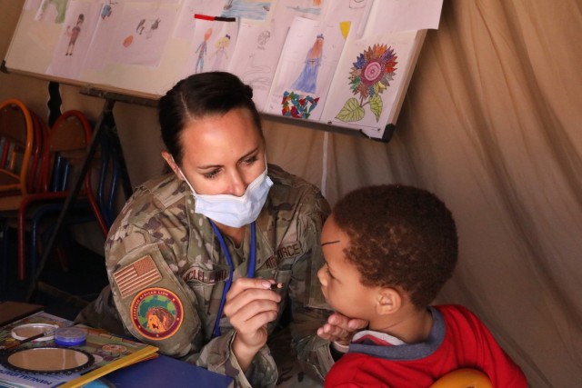 Female soldier seated at desk, holds paintbrush to paint child&#39;s face.