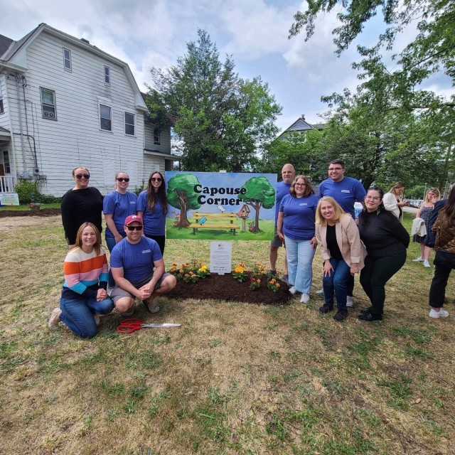 A group of people pose for a photo around a &#34;Capouse Corner&#34; sign and flower bed.