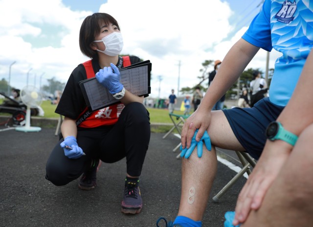 Saho Hirai, a paramedic student with the Kokushikan University Medical Team, listens to a runner explain his ailment at the first-aid station for the Kanagawa Running Association&#39;s 27th annual Friendship Half-Marathon, held April 16, 2023, in conjunction with the Hawaiian Festival on Sagami General Depot, Japan.