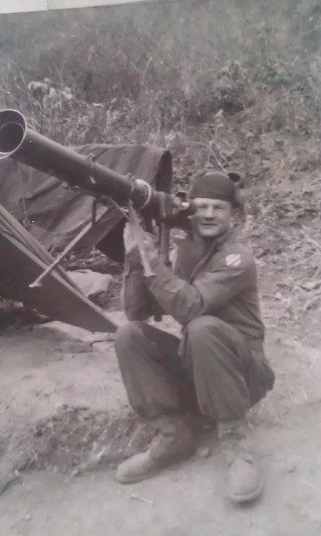 A person sits on the ground and holds a large weapon over their right shoulder. They look at the camera. The photo is black and white.