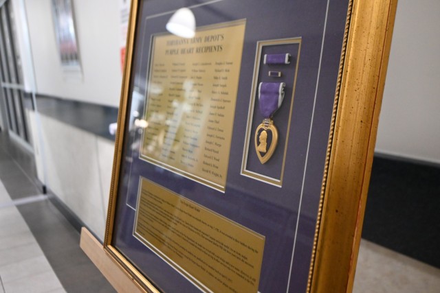 A close-up of a plaque is seen. Names of Purple Heart recipients, a Purple Heart and a history of the award are set on a purple background. A gold frame surrounds the plaque.
