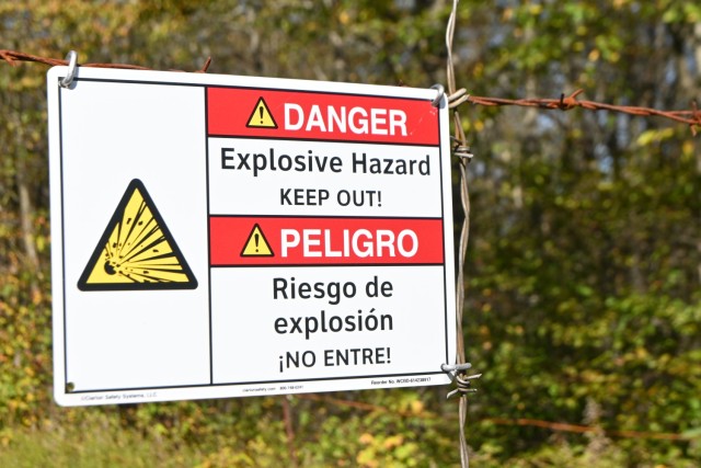 A danger sign is seen hanging on a barbed wire fence in the foreground. The sign has an icon and English and Spanish text warning of explosive hazards. The blurry background contains trees.