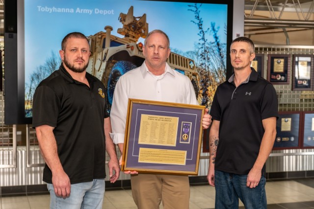 Three people pose for a photo in front of a large tablet with a military vehicle on it. The person at center holds a plaque listing the names of Tobyhanna Army Depot&#39;s Purple Heart recipients.