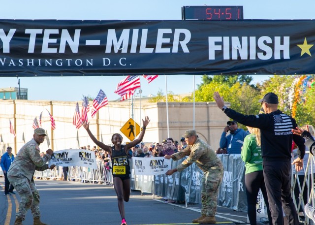 A woman crosses the finish line of the Army Ten-Miler wearing a shirt with the letter AER. Two men in green camouflage uniforms hold the finish line tape that the runner is going through.