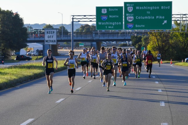 Dozens of runners are running down a highway near the Pentagon with green road signs above them which indicate directions to various local roads.