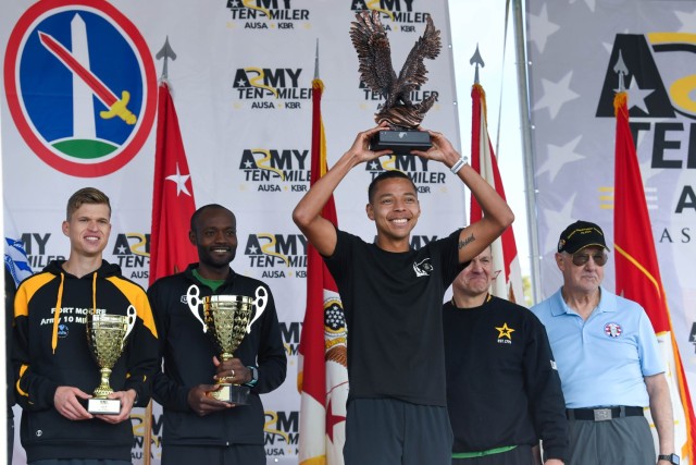A man dressed in a black athletic outfit is holding a bronze trophy above his head which depicts an eagle. Behind him are two men in athletic outfits holding gold trophies that look resemble urns.