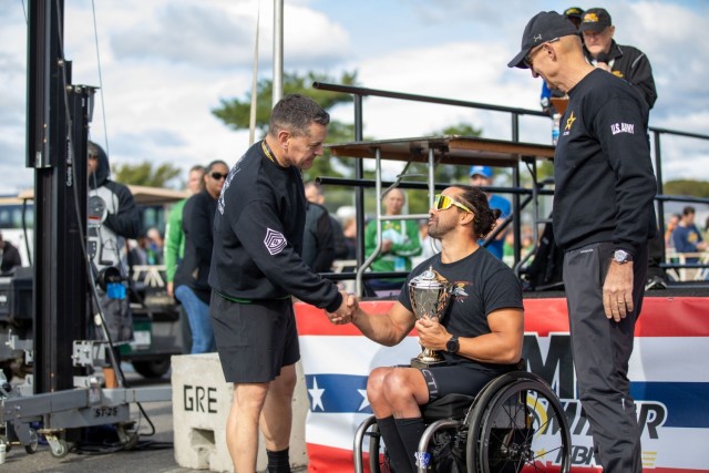 Two men in black athletic outfits are congratulating another man who is in a wheelchair and holding a trophy.