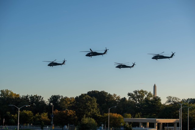 Four helicopters are flying above trees and a parking lot near the Pentagon with the Washington Monument in the background against a light blue, cloudless sky.
