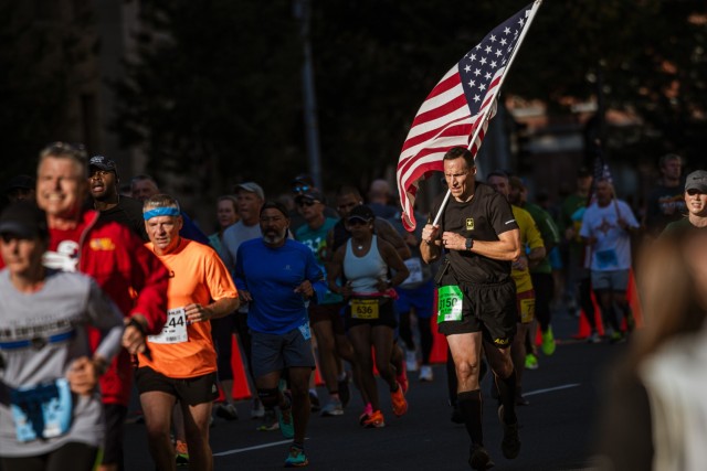 A man wearing black athletic clothes and is carrying the U.S. flag while running in the race with dozens of other runners around him.