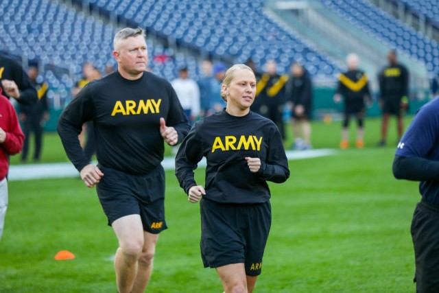 A man and a woman are jogging on green grass. Both are wearing black workout gear that has the word &#34;Army&#34; in large yellow letters.