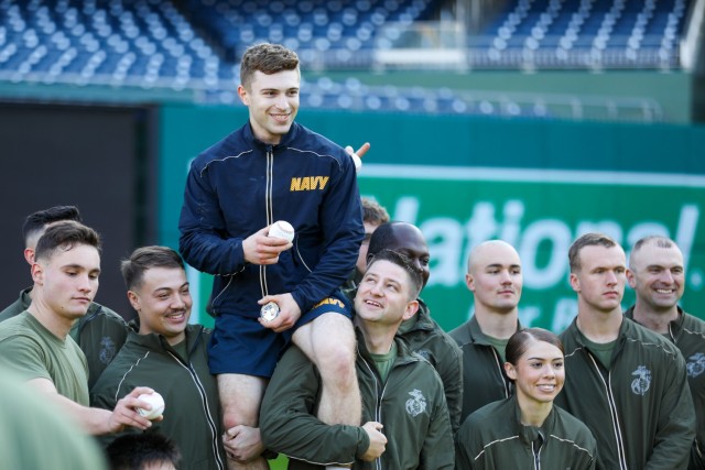 Service members in green U.S. Marine Corps workout gear carry a member dressed in blue U.S. Navy workout gear on their shoulders.