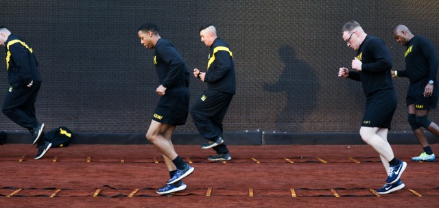 Five service members wearing Army workout gear are jogging though an obstacle course that looks like ladders laying on the ground.