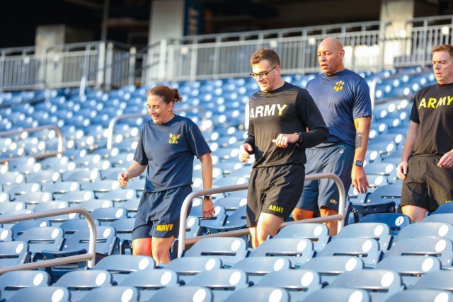 Four service members in workout gear are running down the steps of a baseball stadium. Around them are empty blue seats where the fans would normally sit.