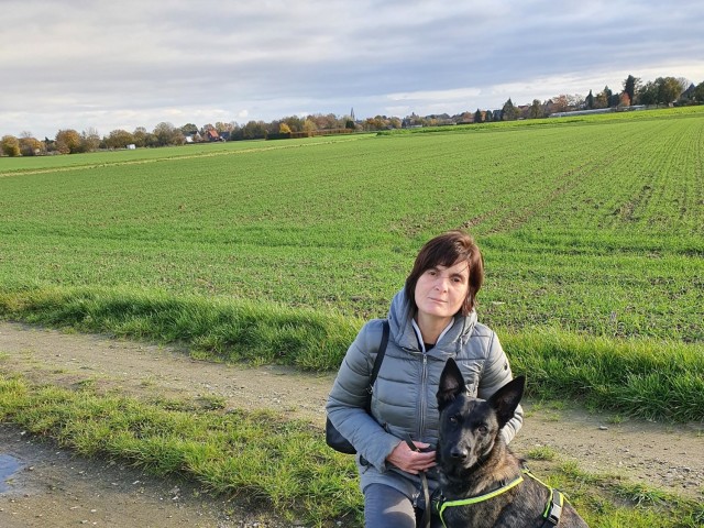 A woman sits next to a dog with a open field behind them. 