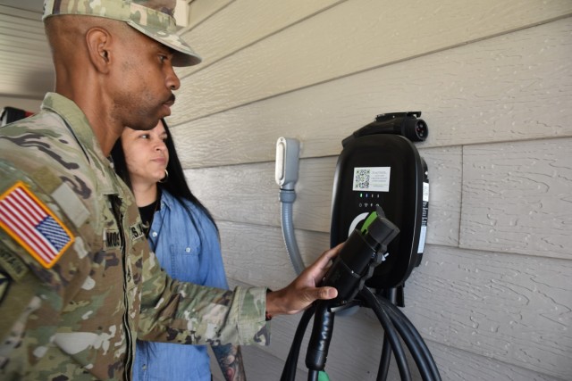 A man in an operational combat uniform holds the cable to an electric vehicle charger while a woman in a denim collared shirt looks on. 