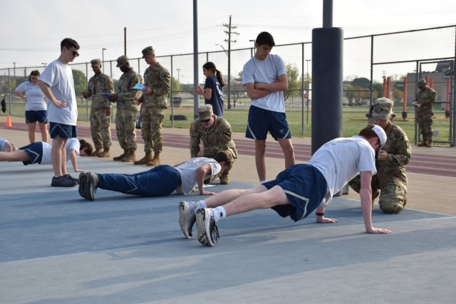 Five men in operational combat uniforms look on at four people in Air Force physical training uniforms completing pushups. 