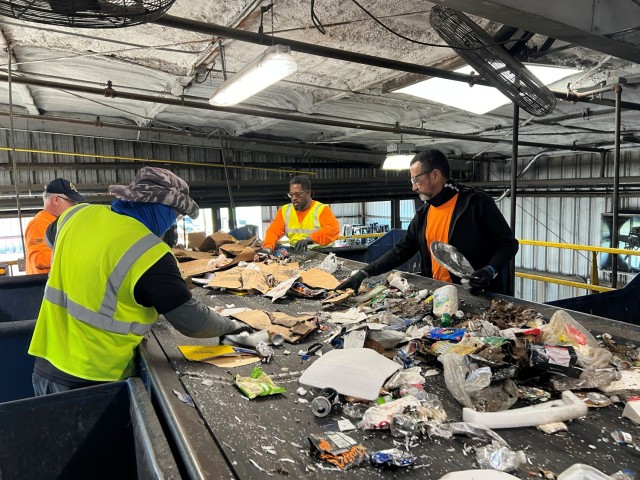 Four men sort through recyclable material on a wide conveyor belt. 