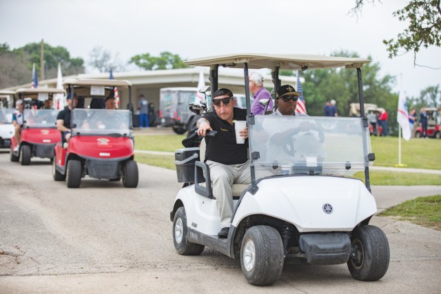 Two men sit in a golf cart, one driving and the other pointing his right pointer finger directly at the camera.