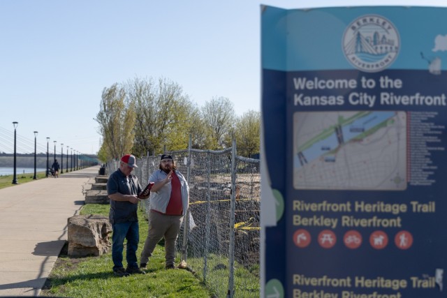 Two men stand in front of a fence with a river behind them and a construction site behind them.
