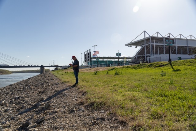 Man standing on bank of river with bridge and stadium in the distance