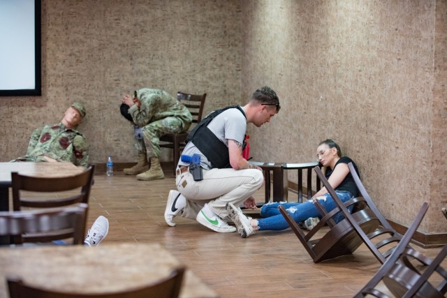 A man crouches down on one knee in front of a woman sitting against the wall on the ground, one leg out and the other crossed underneath. A man ducks his head while sitting in a chair behind them.