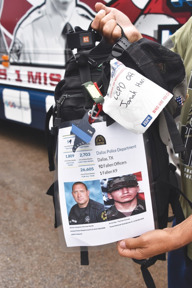 A hand holds a black backpack that has a laminated piece of paper attached to it on a carabiner, along with navy blue keychain in the shape of Texas.