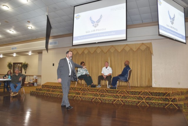 A man stands in an open area, talking, one hand gesturing toward three people sitting and facing each other on a small carpeted stage.