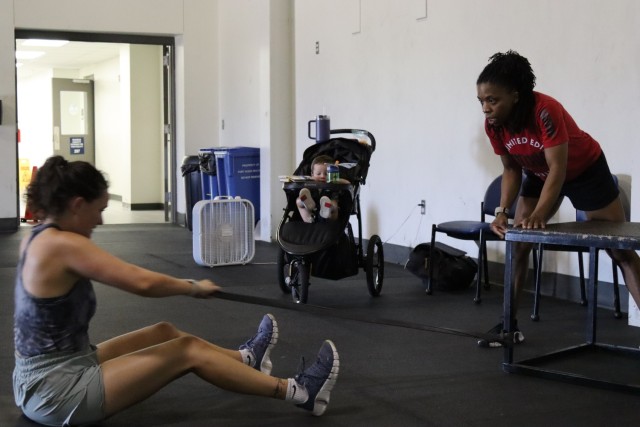 A woman sitting on the floor pulls a resistance band as another woman and a baby boy in a stroller looks on.