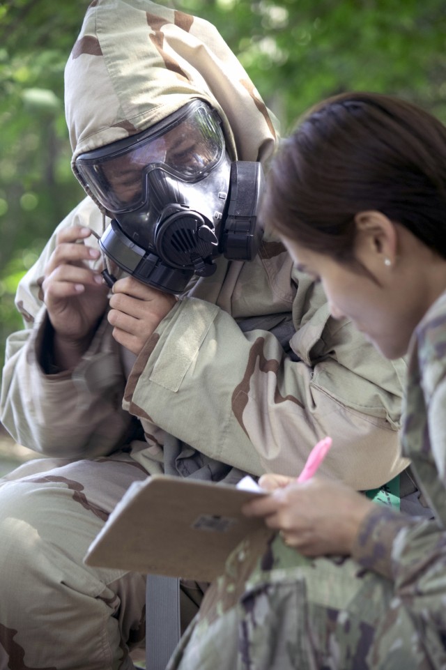 Cadet Rosalyn Page secures the hood of her protective hood over her gas mask as her cadre instructor, Spc. Ye Kim, 68th Chemical, Biological, Radiological, Nuclear, 2nd Battalion, Fort Cavazos, Texas, grades her proficiency on putting on Mission Oriented Protective Posture gear in a timely fashion at Cadet Summer Training on Fort Knox, Kentucky, July 19, 2024. She learned land navigation, first aid, basic rifle marksmanship, hand grenade, and CBRN.