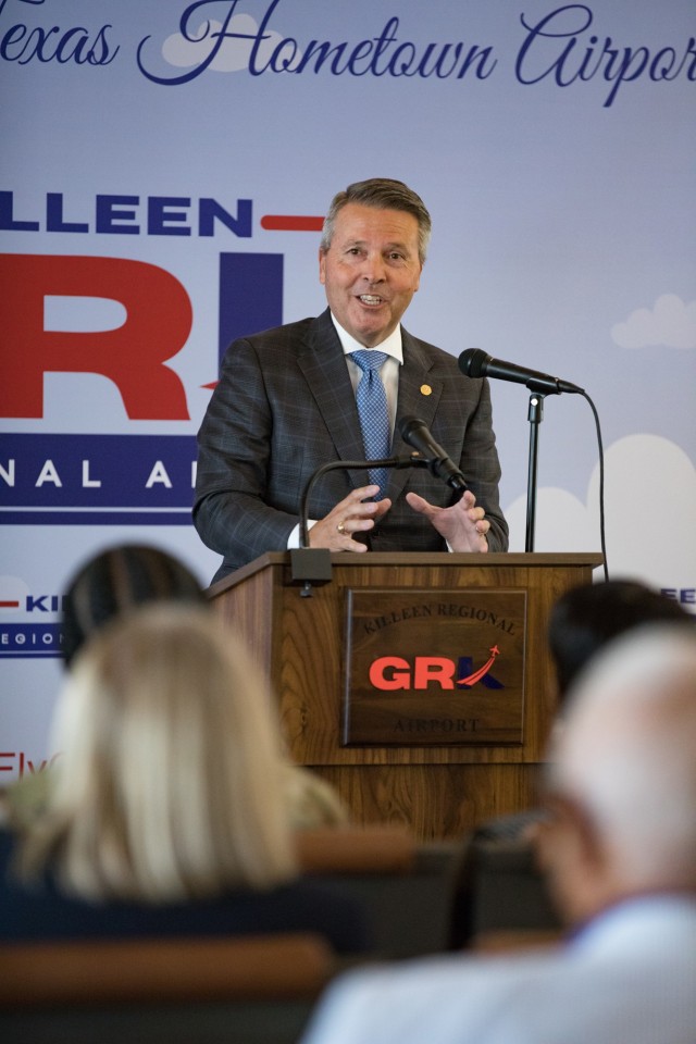 A man standing behind a wooden podium with a mic attached to the top and another sitting on the podium from a stand talks, gesturing with his hands.