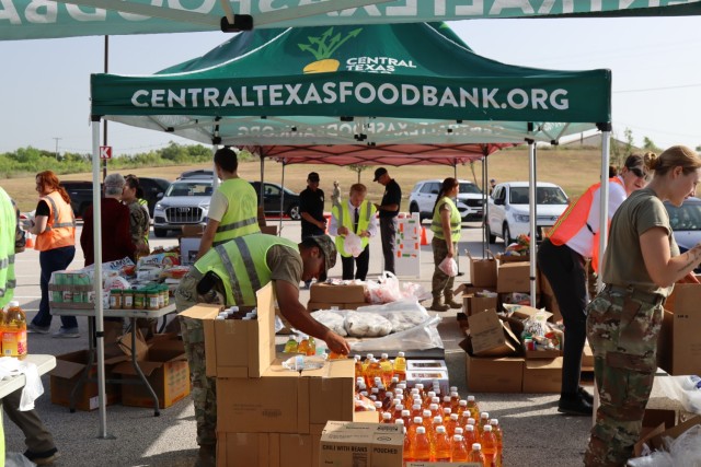 People stand under tents, handling boxes containing various packaged beverages and food.