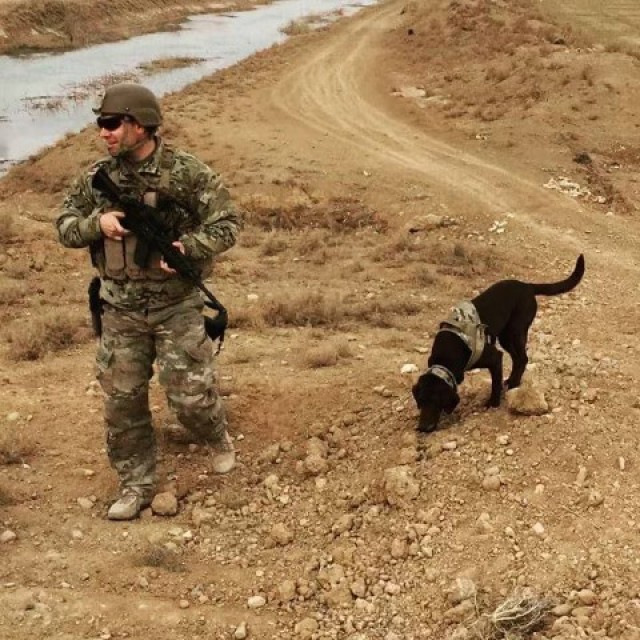 Um homem com uniforme do Exército dos EUA segurando uma arma longa caminha na terra enquanto um cachorro com o focinho no chão o segue.