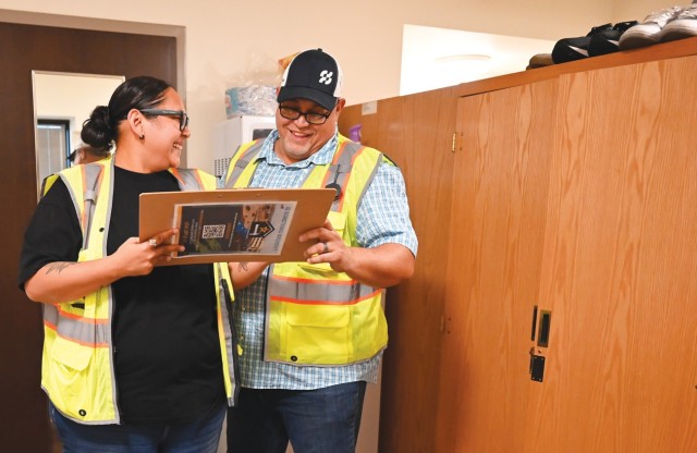 Two people, a man and a woman, hold a clipboard between them while standing and smiling, the man looking at the clipboard and the woman looking at the man.